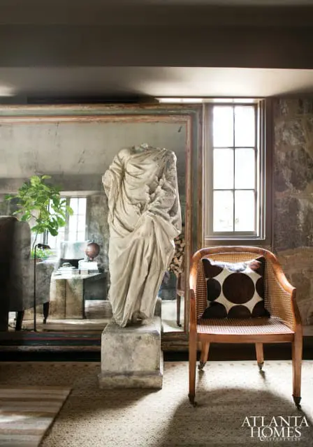 Living room with a stone wall, a large antique mirror reflecting a classical sculpture, and a cane-backed chair with a patterned pillow.