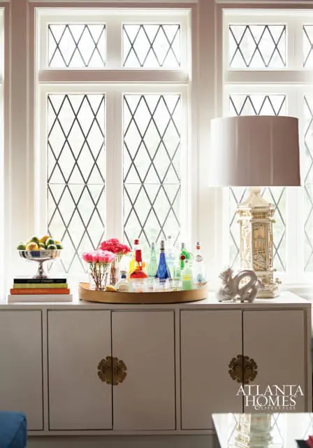 White cabinet with brass hardware below a window with diamond panes, displaying a bar tray with bottles and glasses, a fruit bowl, books, and a decorative lamp and dragon figurine.