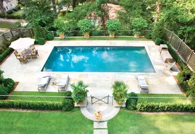 Rectangular swimming pool with turquoise water surrounded by a stone patio, lounge chairs, dining set with umbrella, manicured hedges, and lush trees.