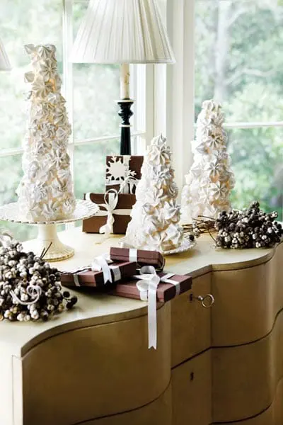 Three meringue trees decorated with white flowers sit on a dresser next to wrapped gifts and decorative branches.