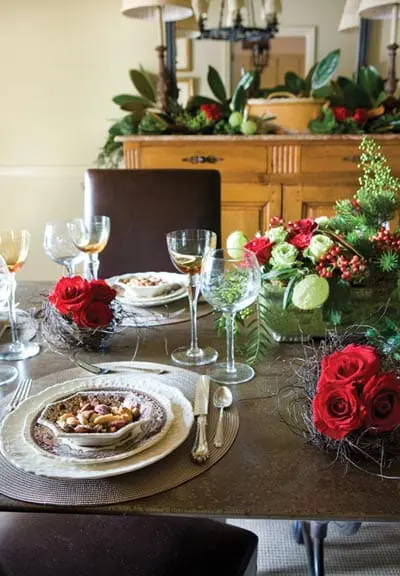 Dining table set with white plates, silverware, wine glasses, and a centerpiece of red roses and greenery. A wooden buffet with a mirror and lamps is visible in the background.