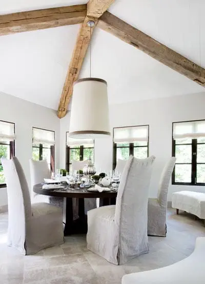 Dining room with a round dark wood table set for a meal, surrounded by six upholstered chairs in light gray slipcovers. Exposed wooden beams cross the white vaulted ceiling above a large drum pendant light.