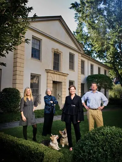 Four people and two corgi dogs pose in front of a light-colored house with stone accents and manicured hedges.