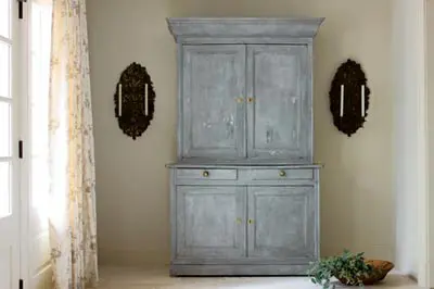 Hallway with a distressed grey hutch, ornate sconces with candles, and a sheer floral curtain next to a white door.