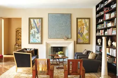 Living room with a fireplace, flanked by artwork and a tall bookshelf filled with books. Two dark sofas and wooden chairs are arranged around a coffee table.