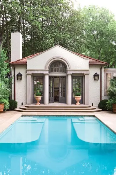 Mediterranean-style pool house with arched entryway, columns, and double doors, set beside a bright blue swimming pool with stone coping.