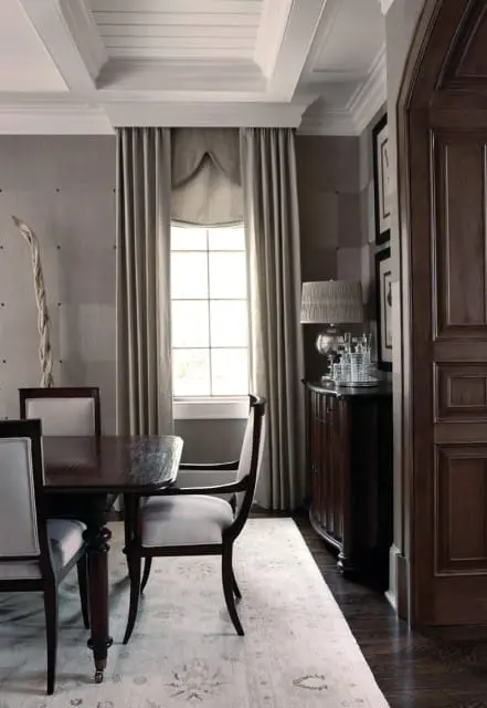 Dining room with dark wood table and chairs upholstered in cream fabric, flanked by grey textured walls and floor-to-ceiling curtains. A dark wood cabinet with a lamp and crystal decanters sits near a window.