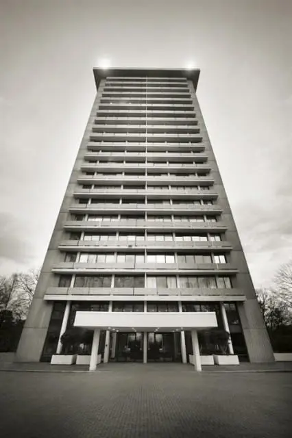 Modernist concrete high-rise building with balconies and large windows, viewed from a low angle on a paved walkway.