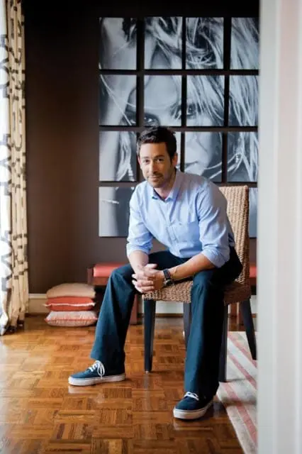 Man in light blue button-down shirt and dark pants sitting on a wicker chair in a room with dark brown walls, a large grid of black and white portraits, and parquet flooring.