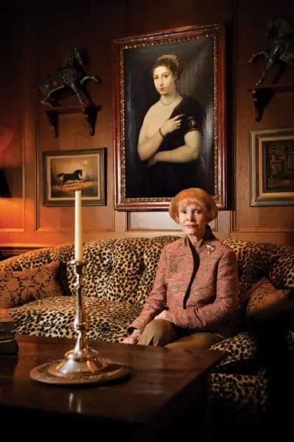 Woman sitting on a leopard print sofa in a room with wood paneled walls, a portrait of a woman above, and decorative horse sculptures.