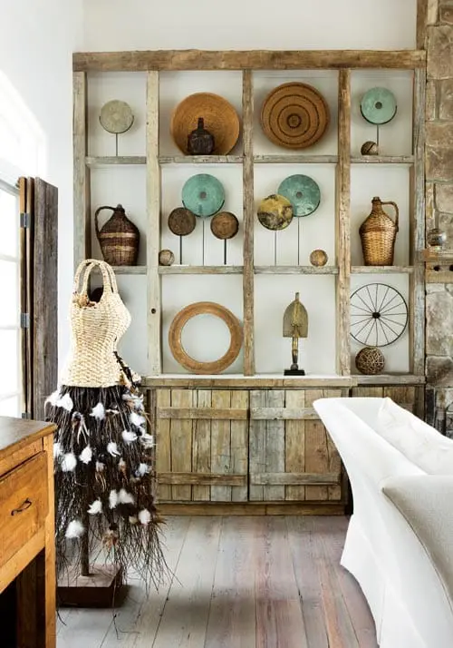 Rustic wooden shelving unit filled with decorative baskets, woven plates, and various art objects. A mannequin form draped with natural materials stands to the left.