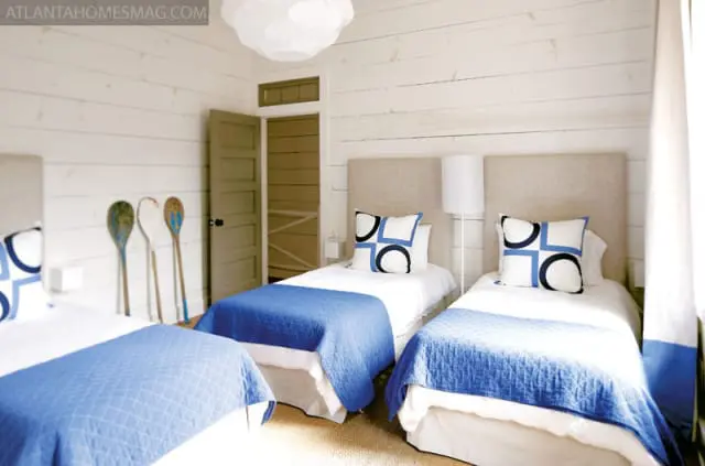 Bedroom with three twin beds, each with a blue quilted coverlet and a neutral upholstered headboard. Walls are white shiplap. Decorative oars lean against the wall.
