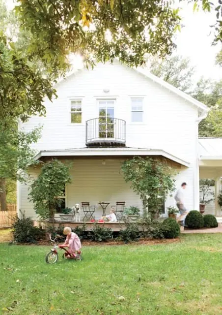 White farmhouse with a covered porch, balcony, and a child riding a tricycle on the lawn.