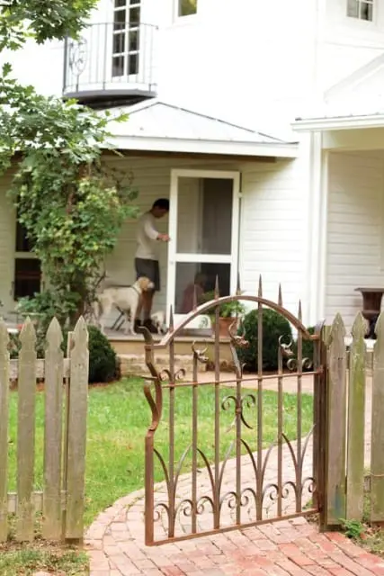 Rustic metal gate and picket fence in front of a white house with a man and two dogs on the porch.