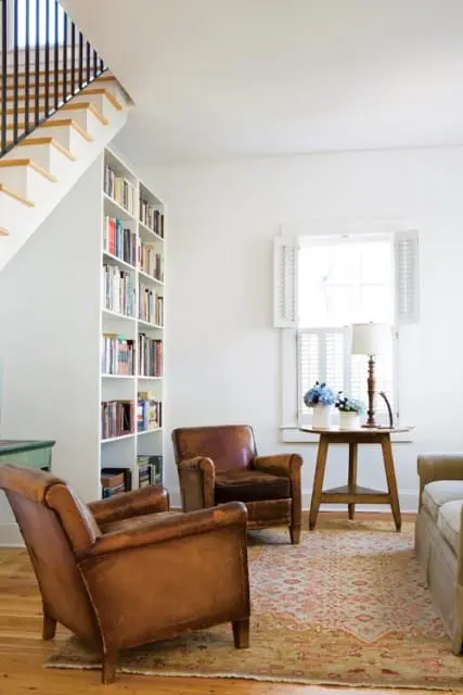 Living room with white walls, a tall white bookshelf filled with books, two brown leather armchairs, and a wooden side table with a lamp and flowers next to a window with white shutters.