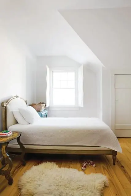 Bedroom with white walls, a white quilted bed, a gold ornate headboard, a wooden side table, a fluffy white rug, and a white paneled door.