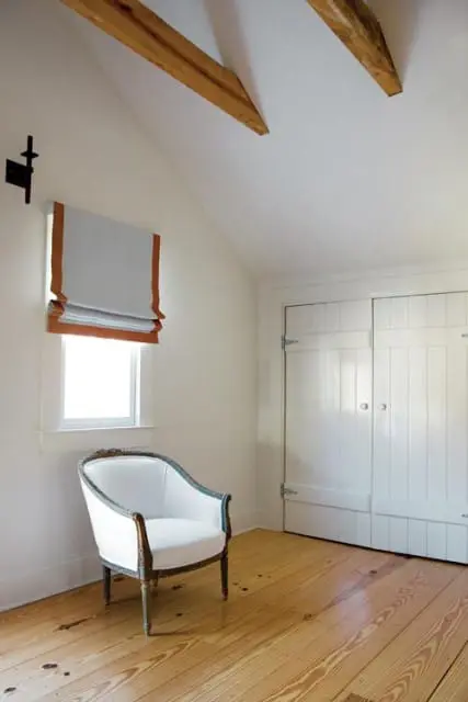 Attic room with white walls, exposed wooden beams, a white armchair with carved wood trim, a window with a Roman shade, and white paneled closet doors.