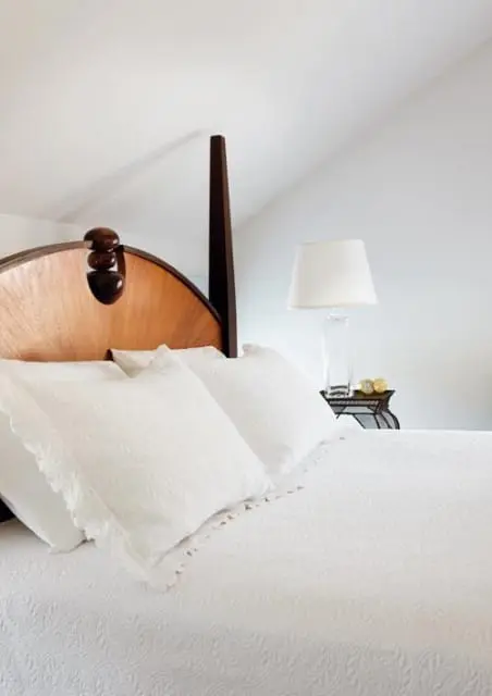 Bedroom with white bedding, decorative pillows, and a wooden headboard with carved details. A white lampshade sits on a glass lamp base on a side table.