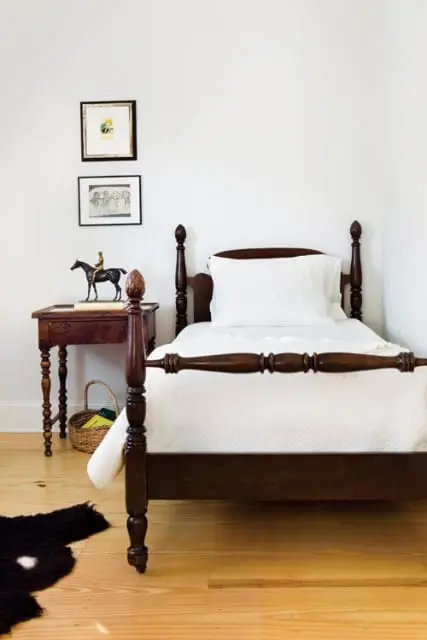 Dark wood four-poster bed with white quilted bedding and pillow. A wooden nightstand with artwork and a horse figurine sits beside the bed.
