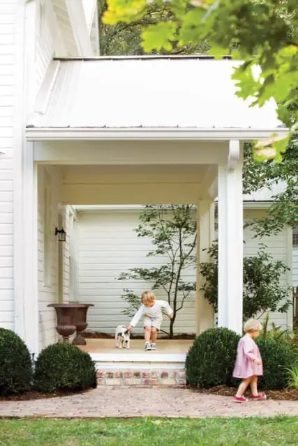 White clapboard house with a covered porch, brick walkway, and manicured boxwood bushes. A young boy pets a dog on the porch while a young girl walks by.
