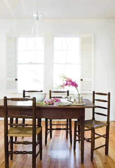 Dining room with dark wood table and chairs with woven seats, set with a white bowl and pink orchids under two shuttered windows.