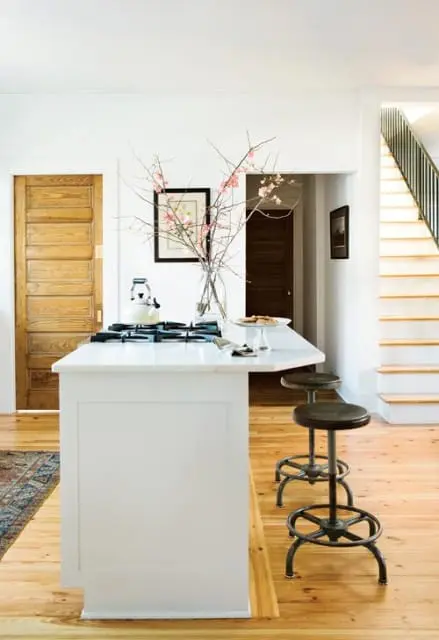 White kitchen island with marble countertop, gas range, and vase of pink blossoms. Two industrial stools sit at the island. Wooden doors and a staircase are visible in the background.