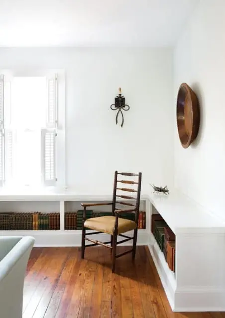 Corner of a room with white walls, built-in white bookshelves filled with books, a wooden chair with a tan seat, and a large wooden bowl mounted on the wall.
