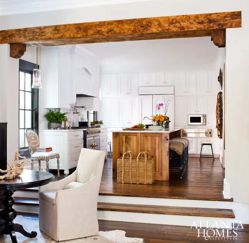 White kitchen with a wooden island, exposed wooden beam, and dark wood floors. A dining area with a round table and chairs is in the foreground.