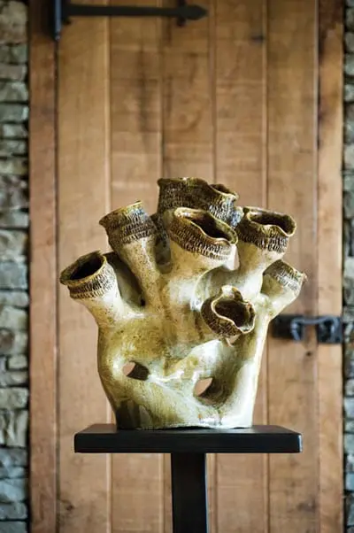 Sculptural ceramic piece with multiple openings and textured rims, displayed on a dark stand against a rustic wooden door and stone wall.