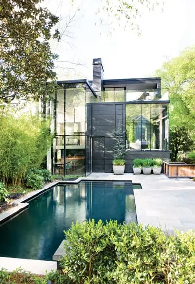 Modern black house with floor-to-ceiling glass windows, dark wood siding, and a dark pool in the backyard surrounded by lush greenery and stone patio.