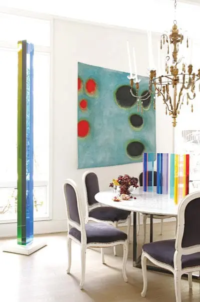 Dining room with marble-topped table, dark upholstered chairs, abstract art, and colorful glass sculptures.