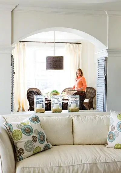 Cream sofa with patterned throw pillows in front of an arched doorway leading to a dining area with wicker chairs and a table set for a meal.