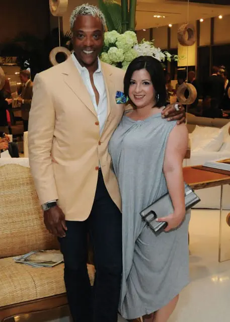 Man with silver hair wearing a peach blazer and white shirt, and a woman wearing a gray dress, posing together indoors.
