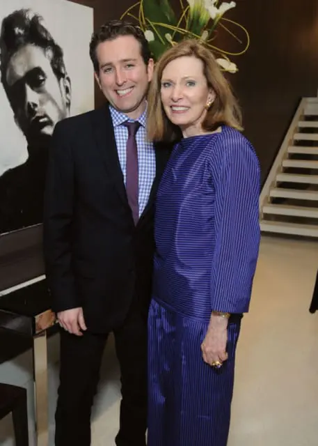 A man in a suit and tie stands next to a woman in a purple striped dress. Behind them is a large black and white portrait and a staircase.