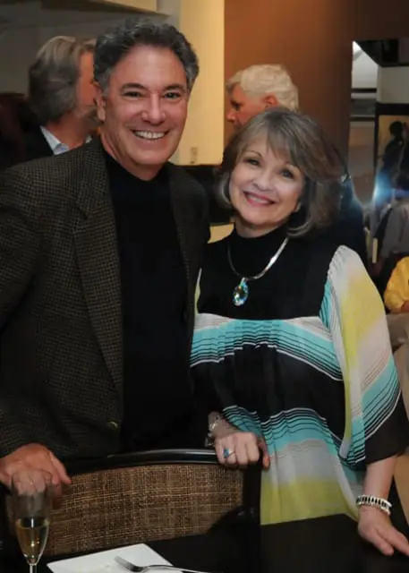 A smiling man in a patterned jacket and a woman in a colorful striped dress stand together, with other people blurred in the background.