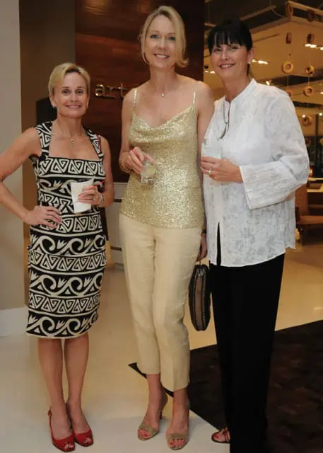 Three women posing for a photo indoors. The woman on the left wears a black and white patterned dress. The woman in the middle wears a gold sequined top and cream pants. The woman on the right wears a white embroidered shirt and black pants.