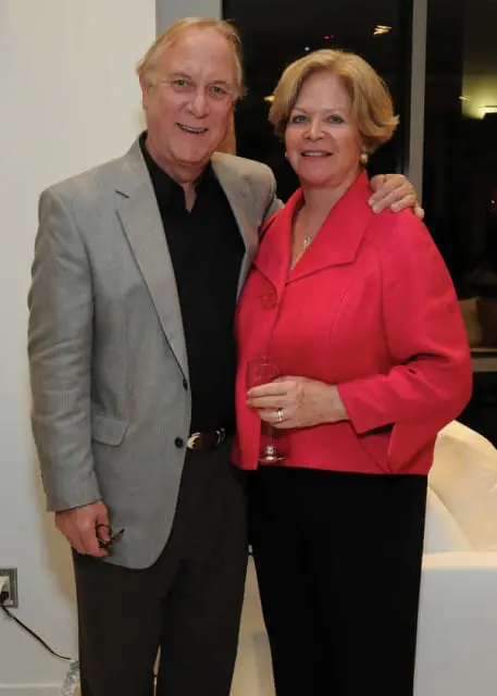 Man in a grey suit jacket and black shirt standing with a woman in a pink jacket. He holds glasses in his left hand.