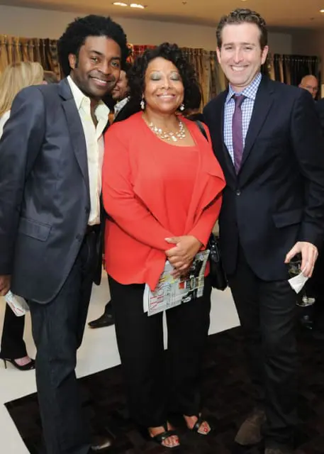 Three people smiling for a photo: a man in a navy suit and light yellow shirt, a woman in a red jacket and black pants, and a man in a navy suit and blue checkered shirt with a purple tie.
