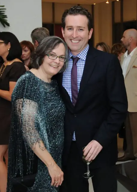 A woman with gray hair and glasses smiles as she stands next to a man in a suit and tie. The man holds a glass of white wine.