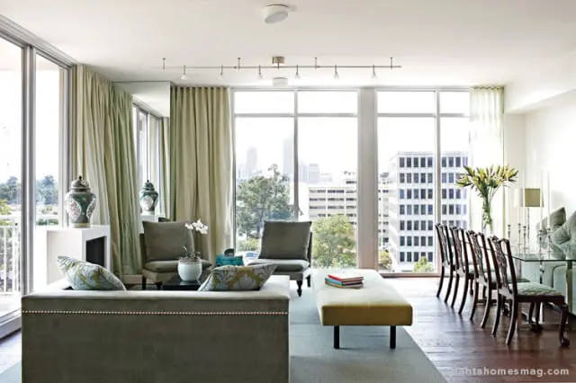 Living and dining area with large windows overlooking a city skyline. Features a green sofa, bench, chairs, and dining set with upholstered seats.