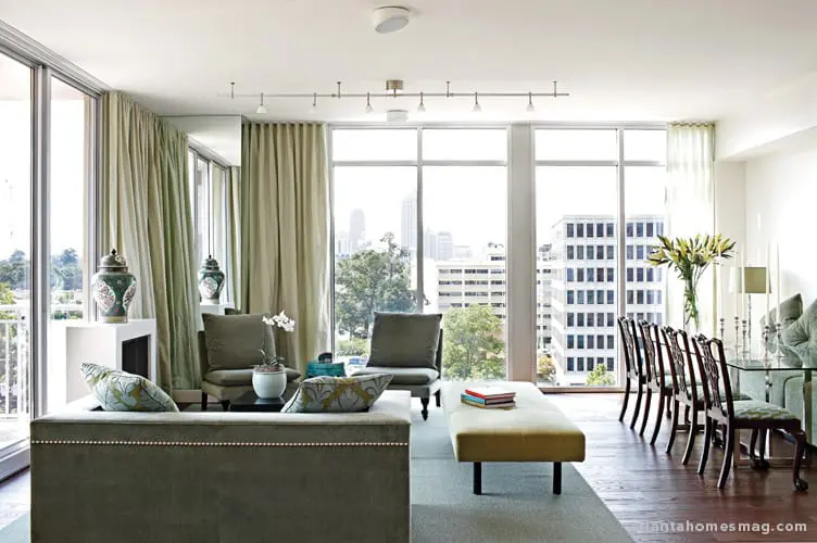 Living and dining area with large windows overlooking a city skyline. Features a green sofa, bench, chairs, and dining set with upholstered seats.