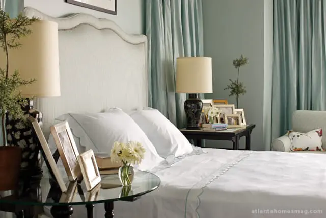 Bedroom with a white upholstered headboard, crisp white bedding with light green trim, and pale blue curtains. A dark lamp sits on a side table with framed photos and flowers.