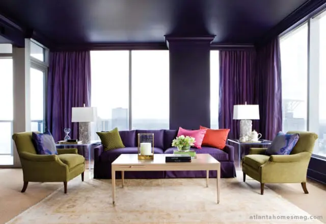 Living room with deep purple walls and ceiling, purple sofa with colorful accent pillows, two olive green armchairs, and a light wood coffee table.