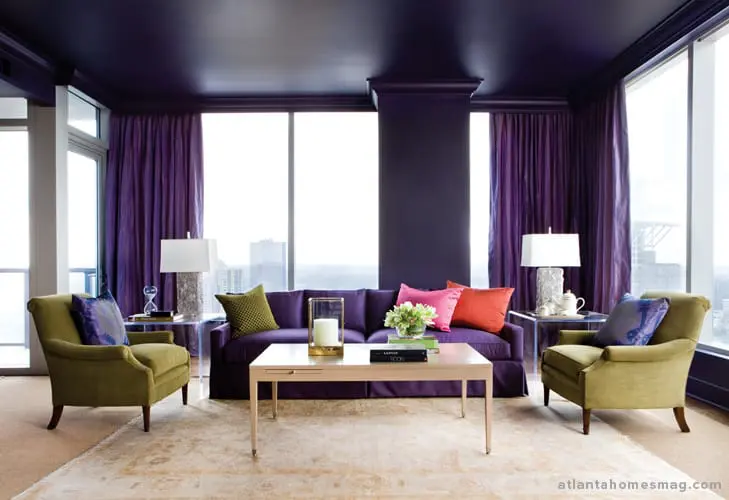 Living room with deep purple walls and ceiling, purple sofa with colorful accent pillows, two olive green armchairs, and a light wood coffee table.