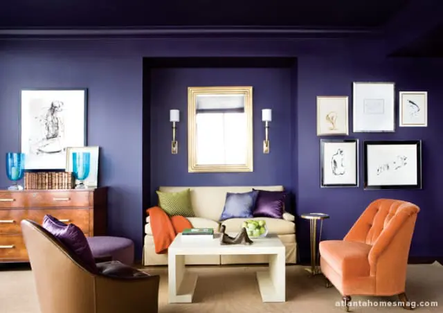 Deep purple living room with a cream sofa, orange armchair, and a white coffee table. Artwork adorns the walls, and a gold-framed mirror hangs above the sofa.