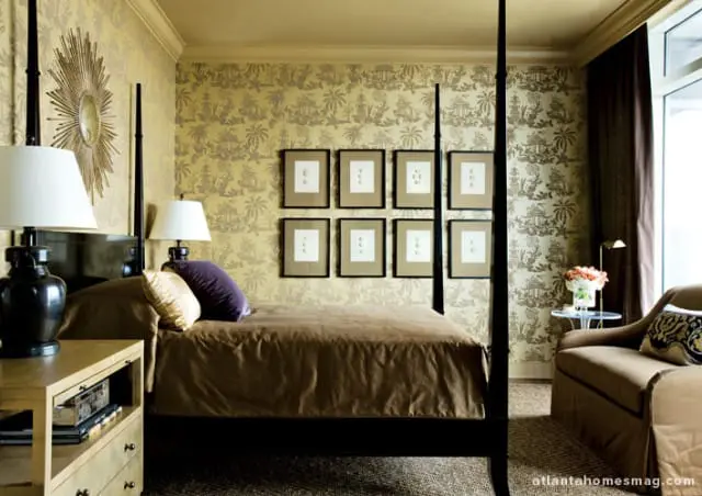 Bedroom with a four-poster bed, gold toile wallpaper, sunburst mirror, and framed botanical prints. A brown duvet covers the bed, with accent pillows.