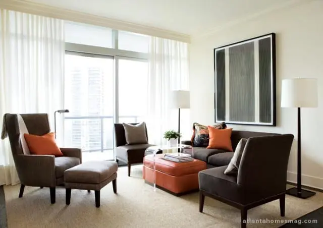 Living room with dark brown upholstered furniture, including a sofa, armchairs, and an ottoman. Orange and patterned throw pillows add pops of color. A large abstract artwork hangs on the wall above the sofa.