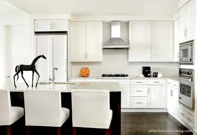 White kitchen with white cabinetry, granite countertops, stainless steel appliances, and a dark wood island with white upholstered stools. A black horse sculpture sits on the island.