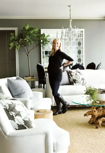 Woman in black dress and boots standing in a living room with white sofas, a large potted plant, and a white chandelier.