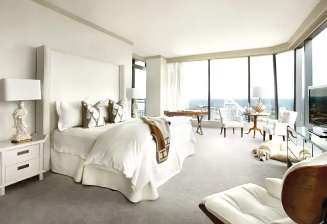 Bedroom with a white upholstered four-poster bed, grey carpet, and large windows overlooking a city skyline. A golden retriever rests on the floor near a mirrored dresser.